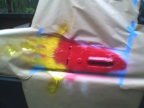 1978 Ford Bronco - A red car door handle is masked and being spray painted with yellow and blue paint, with drips visible.