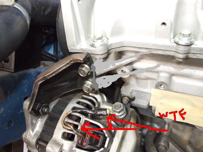 Close-up of car alternator bolted to engine block. Red arrows point to wiring connections. Text 'WTF' is written on tape