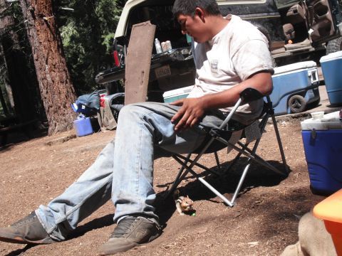 1991 Ford Bronco - A man relaxes in a camping chair while a chipmunk approaches, with coolers and a vehicle nearby in a wooded area.
