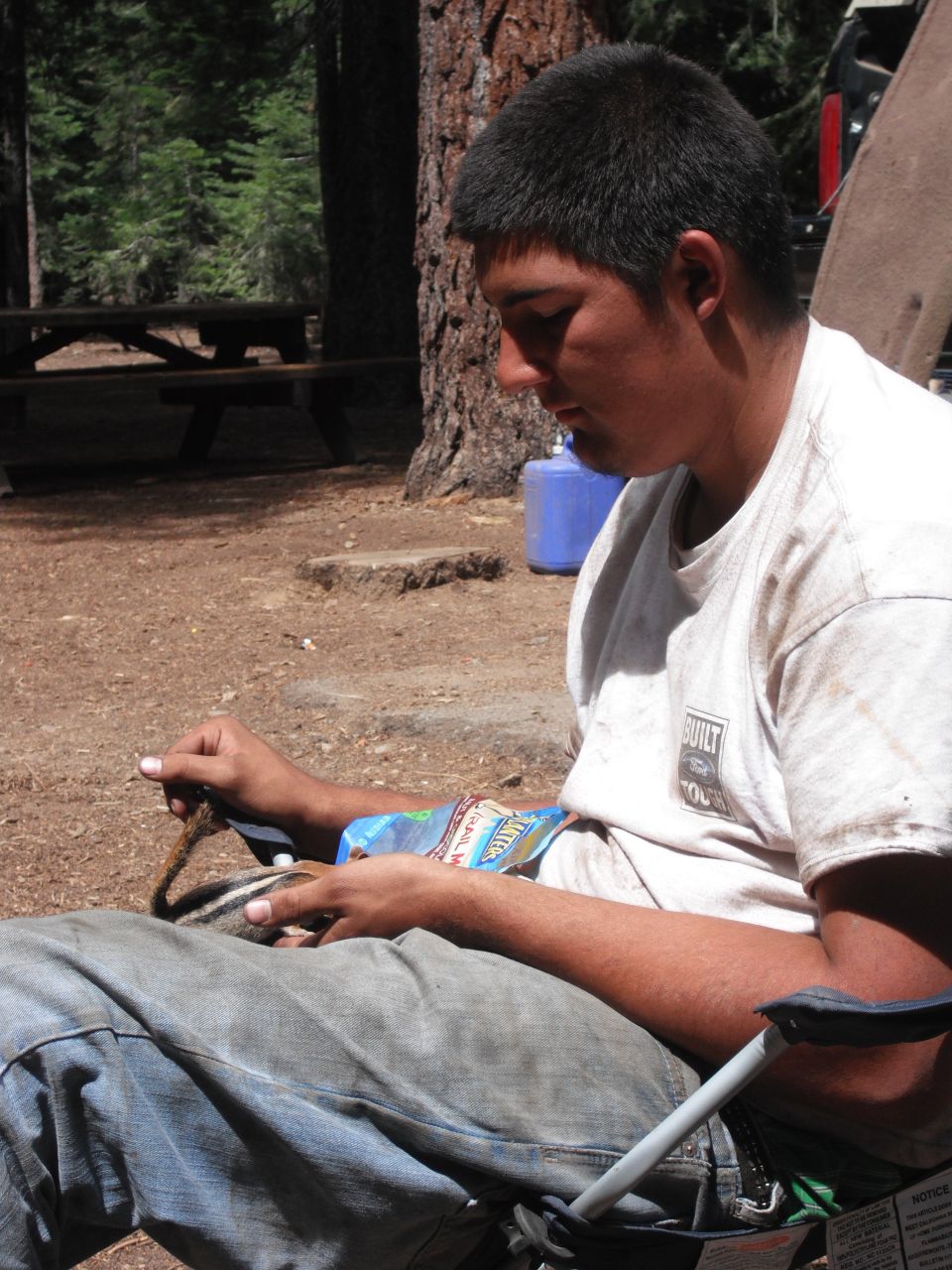 Man in a camping chair feeding a chipmunk from his hand, with a blue water jug and picnic table in the background