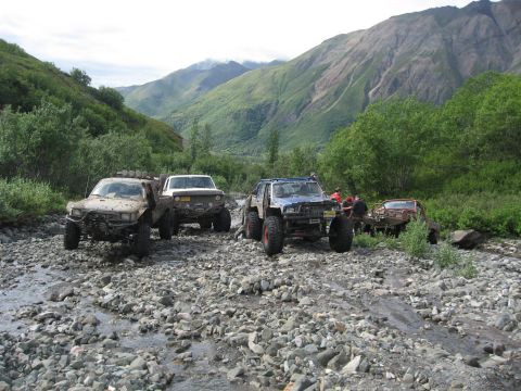 1990 Ford F-250 - Modified trucks are parked in a rocky streambed, surrounded by lush green mountains and trees.