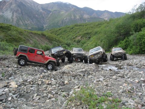 1990 Ford F-250 - A group of off-road vehicles, including a red Jeep Wrangler, traverse a rocky stream bed with a mountainous backdrop.