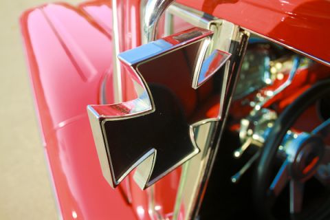 1959 Ford Ranchero - A close-up of a chrome and black cross emblem on the exterior of a red vehicle, with a glimpse of the steering wheel and dashboard inside.