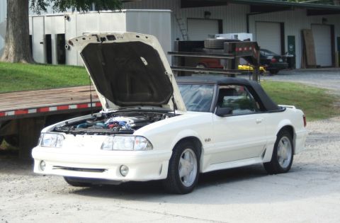 1976 Ford Ranchero - A white convertible Mustang is parked outside a building with its hood open, revealing the engine bay.