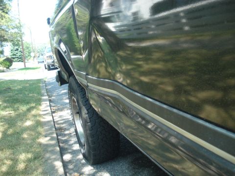 1988 Ford Bronco - A dark green Ford Bronco is parked on the side of a residential street, showing its side profile and tire.