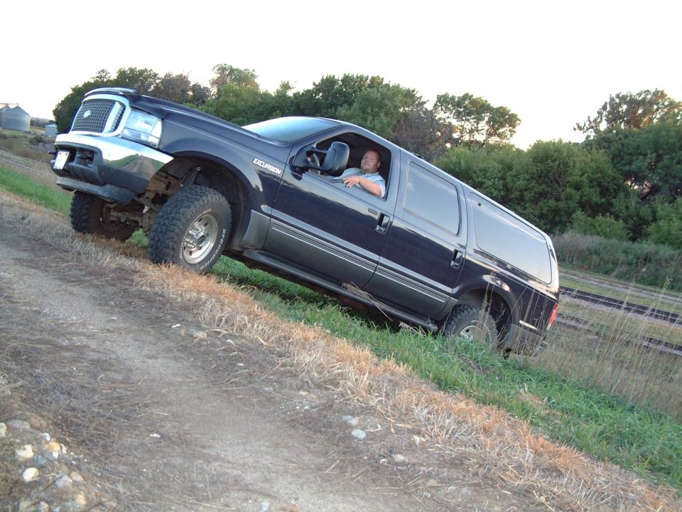 Dark blue Ford Excursion SUV driving on a dirt incline with grass on the side. Driver is visible