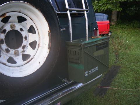 1981 Ford E350 - An ammo can is mounted to the side of a vehicle next to a spare tire, with a red pickup truck visible in the background.