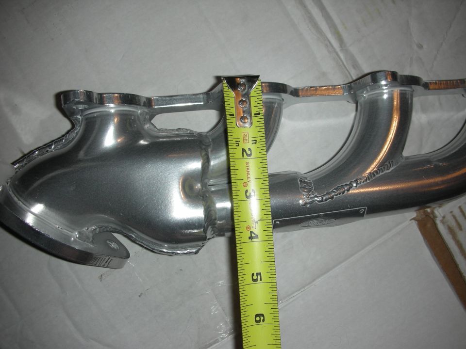 Close-up of an aluminum exhaust manifold with a yellow tape measure showing dimensions
