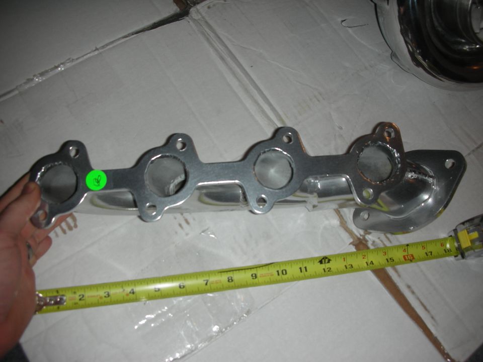 Chrome exhaust manifold with a green sticker and a yellow measuring tape