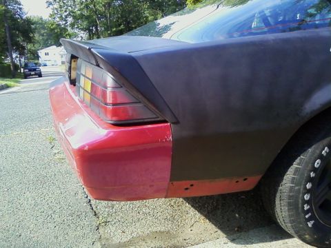 1989 Chevrolet Camaro - The rear of a car shows a red bumper and a black, unfinished quarter panel, with a spoiler on the trunk.