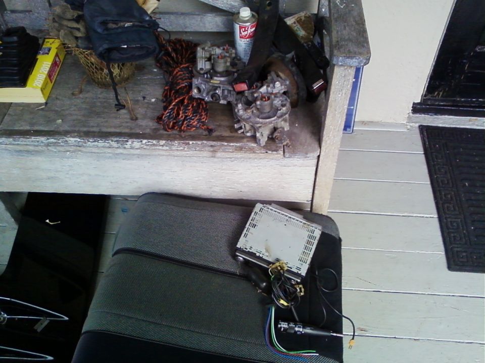 Two carburetors and Sea Foam can on wooden bench; car stereo with wiring on a removed gray seat below