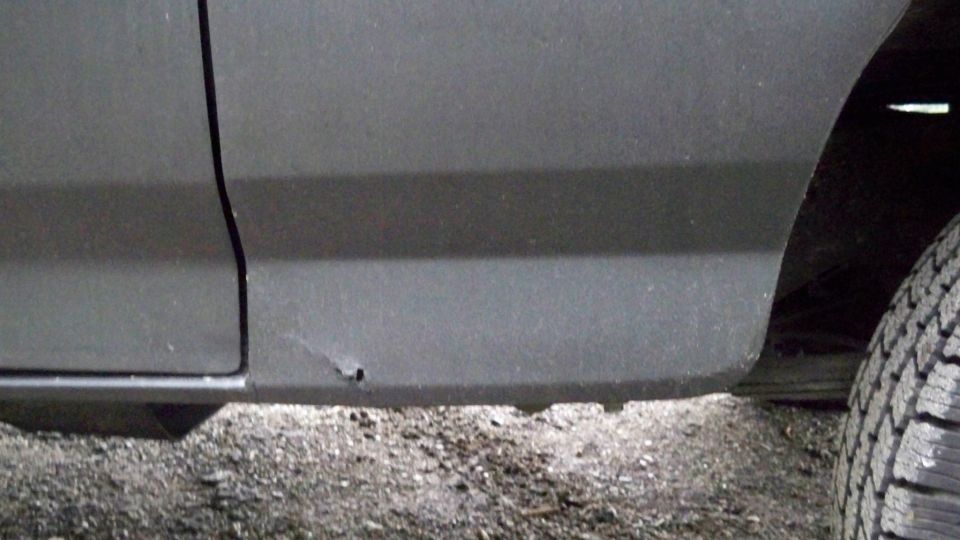Gray vehicle lower body panel with a small rust spot and a tire visible on the right