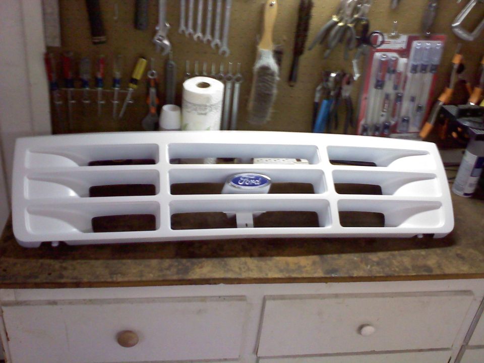 White Ford Bronco grille with blue Ford oval emblem. Tools on pegboard in background