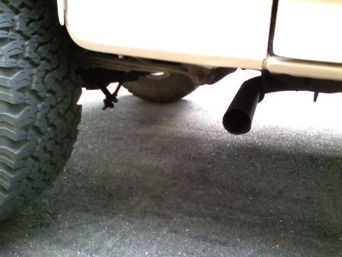 1990 Ford Bronco - A black exhaust pipe extends from the underside of a vehicle, with a large tire in the foreground.