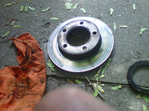 1995 Ford Bronco - A removed vented brake rotor lying face-up on cracked concrete, its friction surface scored and lipped from wear and the stud holes empty.…