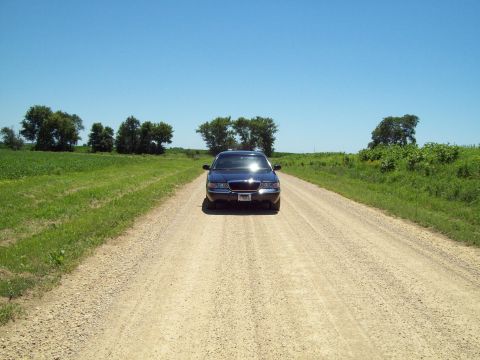 1999 Mercury Grand Marquis - Dark blue Mercury Grand Marquis sedan sitting head-on in the middle of a straight gravel country road, framed by green crop fields and scat…