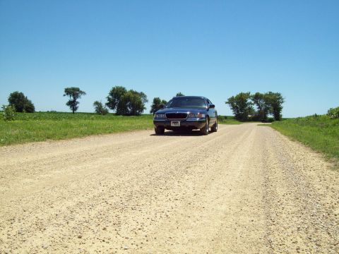 1999 Mercury Grand Marquis - Wide low shot of a dark blue Mercury Grand Marquis parked facing the camera at the edge of a gravel country road, with the coarse gravel su…