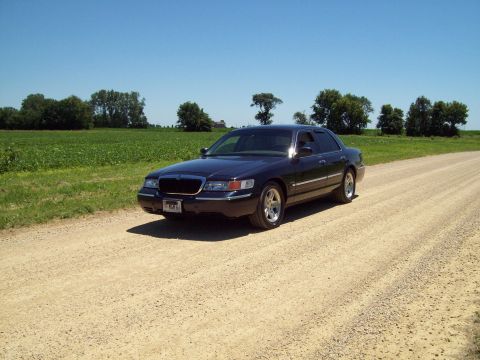 1999 Mercury Grand Marquis - Dark blue Mercury Grand Marquis sedan parked on a gravel road beside a green crop field under clear blue sky, shot from a distant front thr…