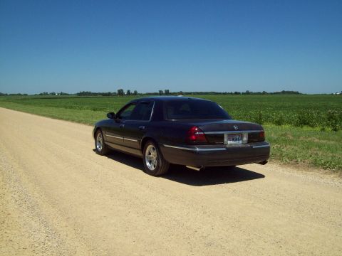 1999 Mercury Grand Marquis - Dark blue Mercury Grand Marquis sedan parked on a gravel farm road, shot from the driver-side rear three-quarter angle with green crop fiel…