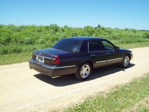 1999 Mercury Grand Marquis - Dark navy blue Mercury Grand Marquis sedan on a gravel road beside a green weedy field under a clear blue sky, shot from the passenger-side…