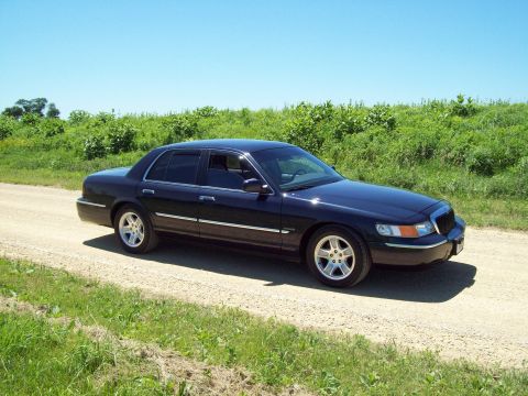 1999 Mercury Grand Marquis - Dark navy blue Mercury Grand Marquis sedan on a gravel country road bordered by tall green weeds under a clear blue sky, shot from a low si…