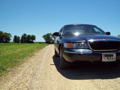1999 Mercury Grand Marquis - Low ground-level shot of the passenger-side front corner of a dark blue Mercury Grand Marquis on a gravel country road, with sun glinting o…
