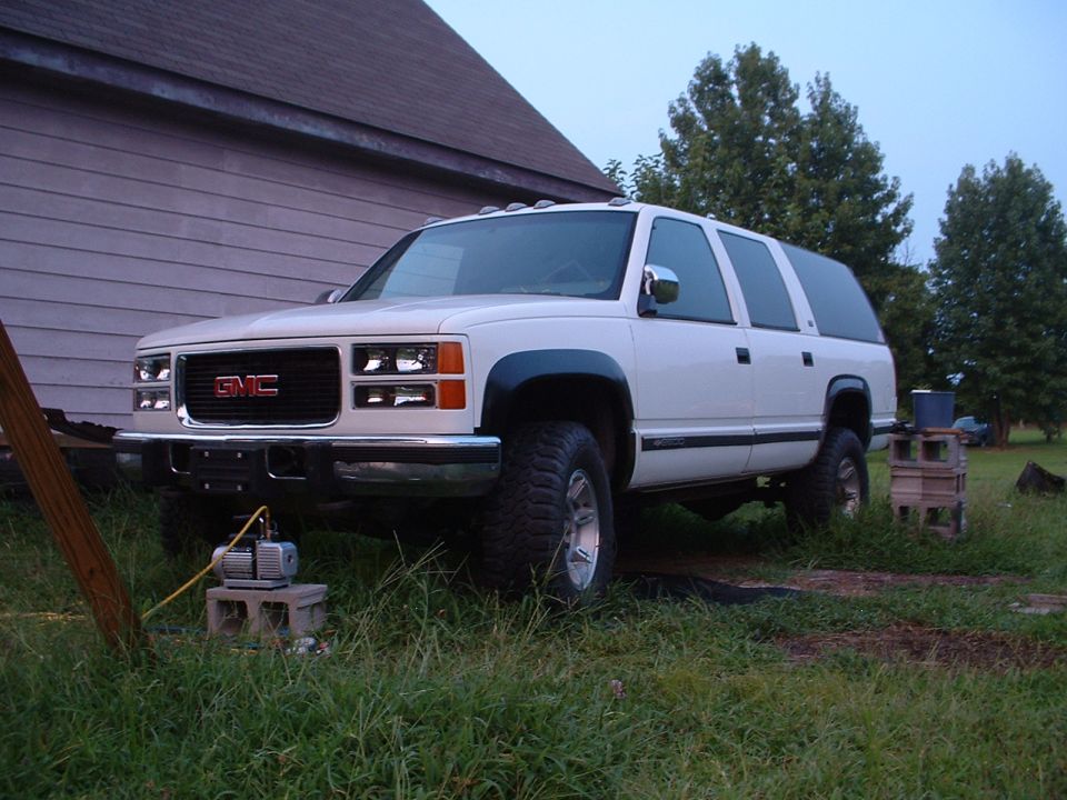 White GMC Suburban parked on grass, front 3/4 view. Visible Supermotors.net logo