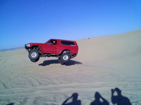 1994 Ford Bronco - A red 1994 Ford Bronco is captured mid-air while jumping over a sand dune under a clear blue sky.