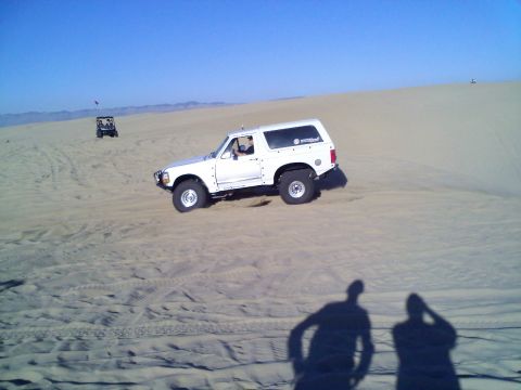 1994 Ford Bronco - A white 1994 Ford Bronco navigates sand dunes under a clear blue sky, with another off-road vehicle visible in the distance.