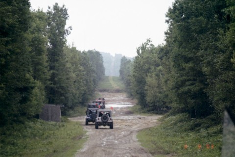1979 Ford Bronco II - Several SUVs navigate a wet, muddy path through a wooded area, kicking up spray.
