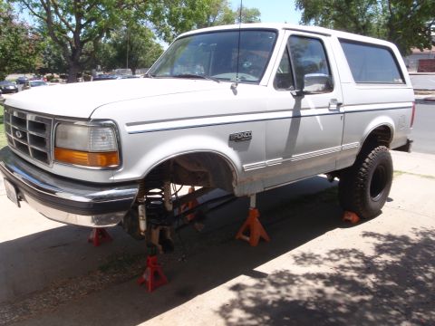 1994 Ford Bronco - A white Ford Bronco XLT with blue pinstriping sits on orange jack stands in a driveway with the driver's front wheel removed, exposing the…