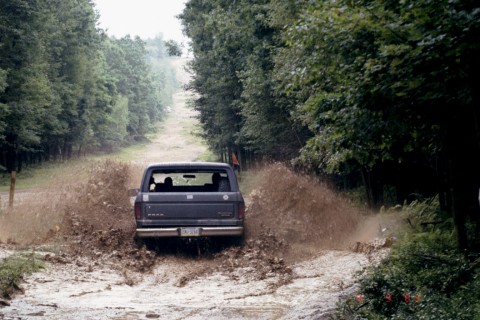 1979 Ford Bronco II - A dark SUV navigates a muddy path, kicking up water and dirt.