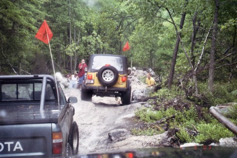 1979 Ford Bronco II - Off-road vehicles tackle a challenging trail with trees and rocks surrounding them.
