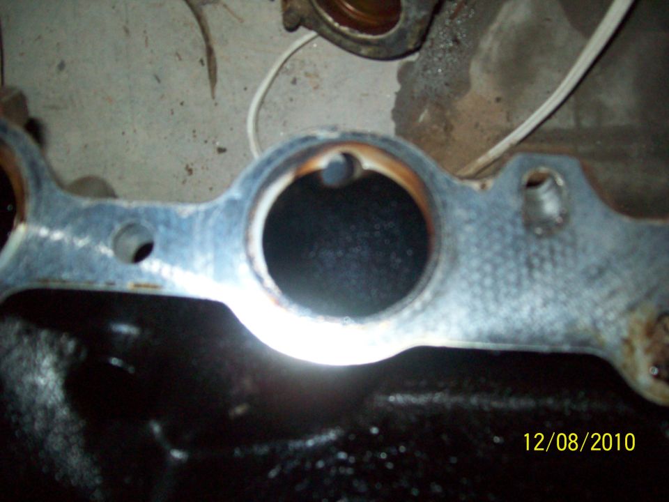 Engine intake manifold with two circular openings, one showing a dark interior. Metal surface shows some rust and wear. Date stamp reads 12/08/2010