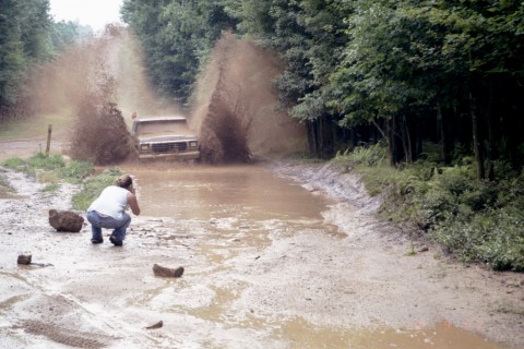 1979 Ford Bronco II - A truck makes a big splash as it drives through a muddy off-road course, with a photographer capturing the action.