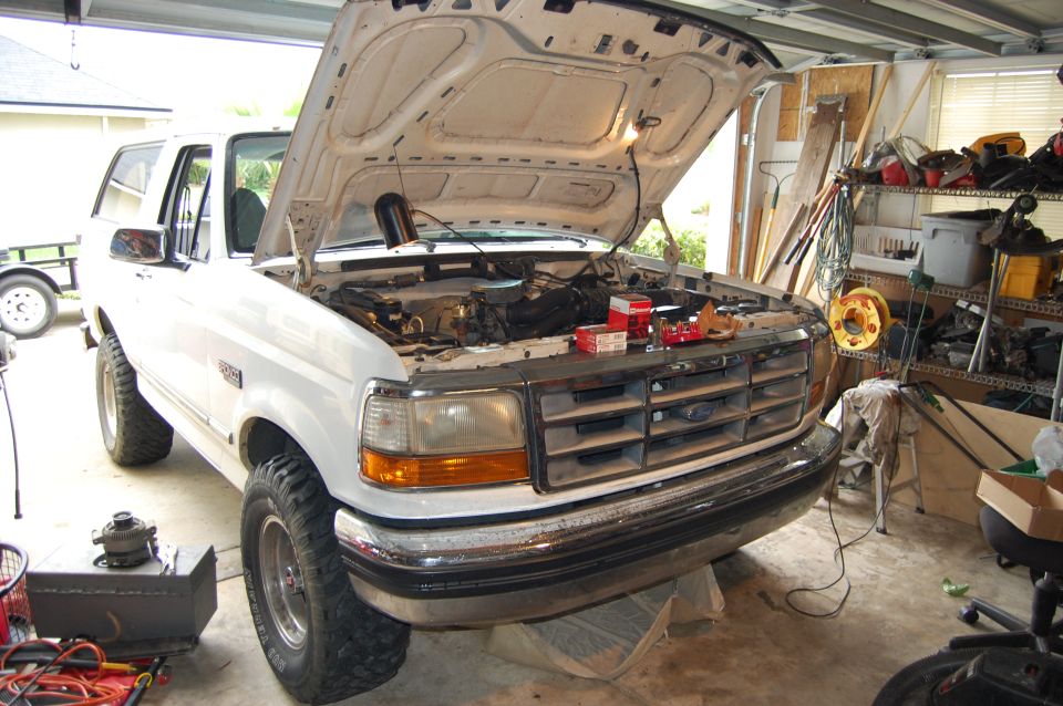 White Ford Bronco in garage, hood up, engine exposed. Parts and tools are visible around the vehicle