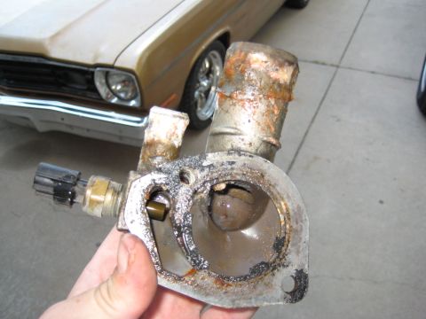 2005 Jeep Wrangler - A hand holds a corroded cast aluminum thermostat housing / coolant outlet removed from an engine, showing rust-stained upper radiator hose…