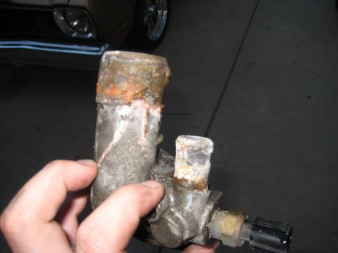 2005 Jeep Wrangler - A hand holds a removed cast aluminum thermostat housing / coolant outlet neck up to the camera, showing heavy orange-brown corrosion and cr…