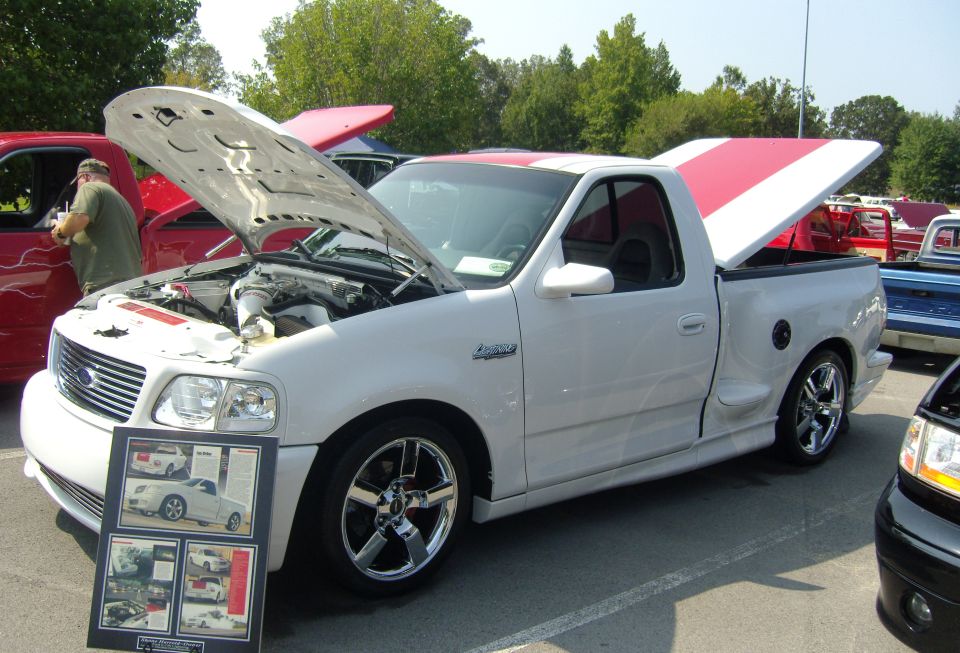 White pickup truck with hood and tonneau cover open, chrome wheels, and a display board in front