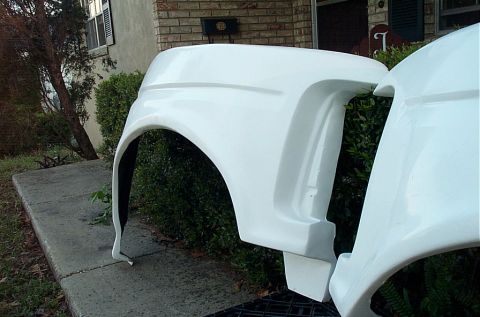 1995 Ford Bronco - Pair of white fiberglass front fenders standing on end against a hedge beside a brick house, photographed from the outside face showing the…