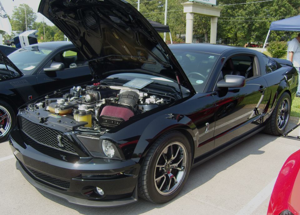 Black Ford Mustang Shelby GT500 with hood open, showing engine. Another Mustang is parked next to it