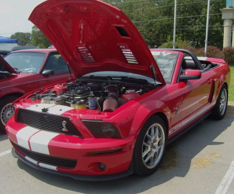 2007 Ford Mustang - A red 2007 Ford Mustang convertible is displayed with its hood open, revealing the engine and prominent white racing stripes.