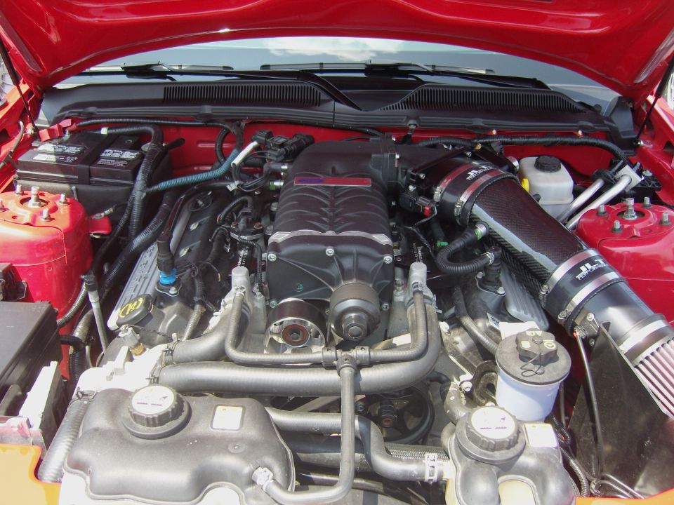 Engine bay with a black supercharger and carbon fiber intake tube. Some hoses and the battery are visible