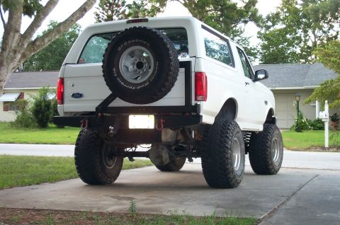 1995 Ford Bronco - Rear view of a white full-size Ford Bronco, lifted on mud tires with a rear bumper-mounted spare tire carrier.