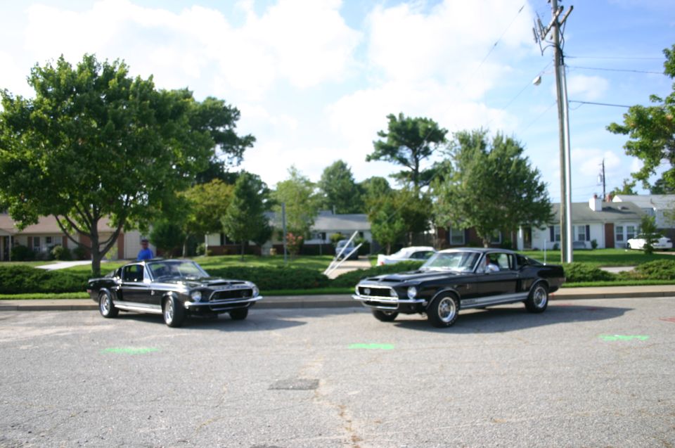 Two black 1968 Shelby GT500 Mustangs parked on asphalt with trees and houses in background