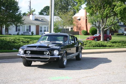 1968 Shelby GT500 - A black 1967 Ford Mustang is seen driving on a street with a red Mustang parked in the background.