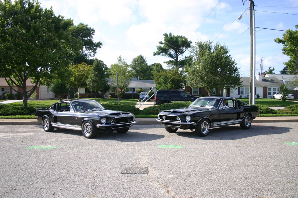 Two black 1968 Ford Mustangs parked side-by-side on asphalt, with houses and trees in the background
