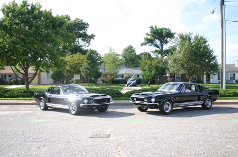 1968 Shelby GT500 - Two classic black Ford Mustangs are parked side-by-side on a suburban street under a bright sky.