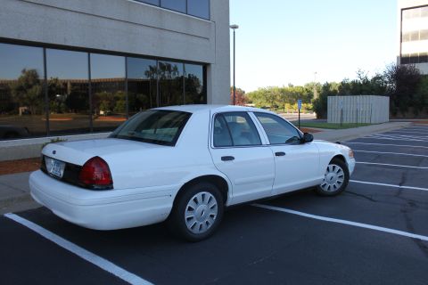 2003 Ford Crown Victoria - A white sedan is parked in a lot next to a modern building.