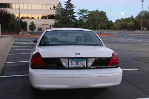 2003 Ford Crown Victoria - Rear view of a white 2003 Ford Crown Victoria parked in an empty lot with a building in the background.